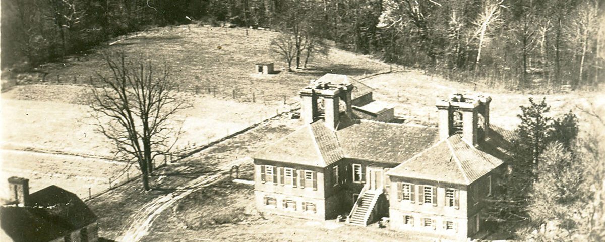 Black and white aerial photograph of landscape featuring open fields edged by woods, with a large brick house