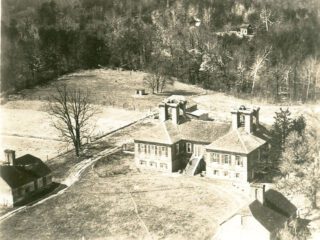 Black and white aerial photograph of landscape featuring open fields edged by woods, with a large brick house