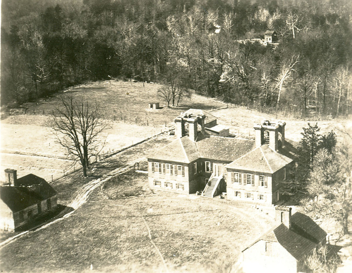 Black and white aerial photograph of landscape featuring open fields edged by woods, with a large brick house