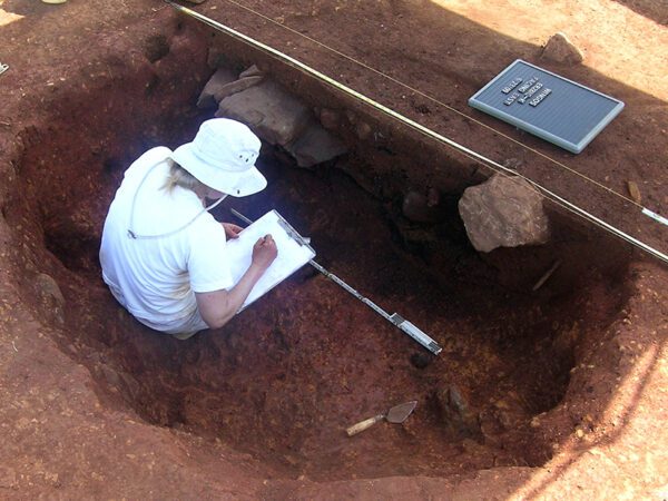 Woman wearing white, inside a red clay excavation unit, drawing a profile