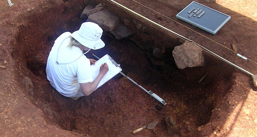 Woman wearing white, inside a red clay excavation unit, drawing a profile