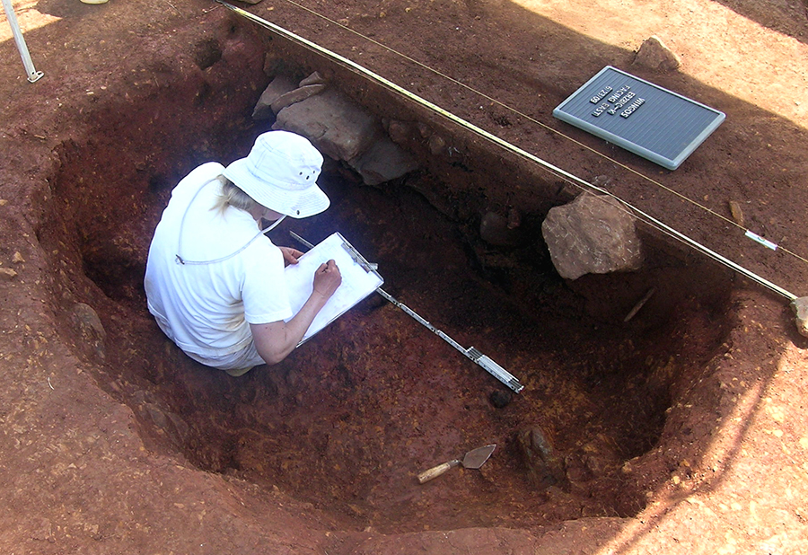 Woman wearing white, inside a red clay excavation unit, drawing a profile
