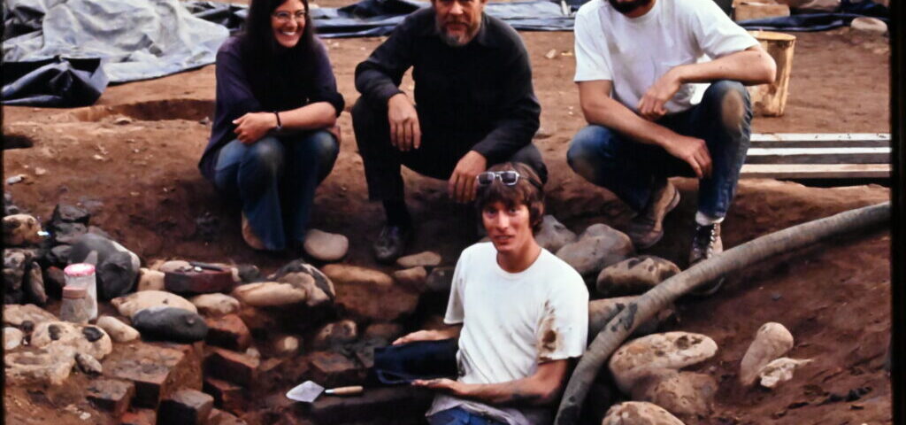 group of four people clustered in a large scale excavation to subsoil with rocks and artifacts protruding