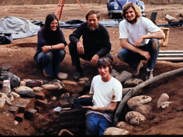 group of four people clustered in a large scale excavation to subsoil with rocks and artifacts protruding