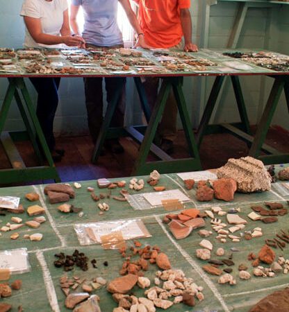 Foreground shows artifacts laid out on plywood tables. In the background, three people are clustered around another table of artifacts.