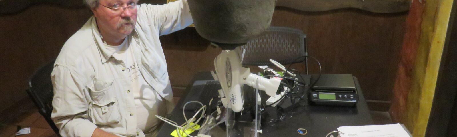 Man seated in museum setting with an antique copper vessel resting on a scientific instrument. Other scientific and recording tools surrounding.