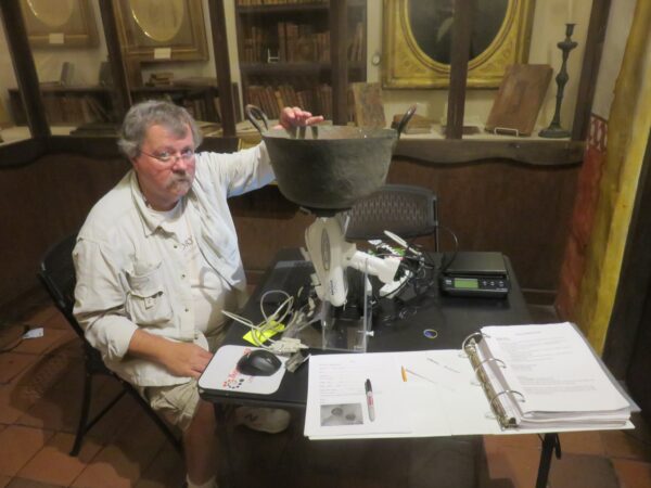 Man seated in museum setting with an antique copper vessel resting on a scientific instrument. Other scientific and recording tools surrounding.