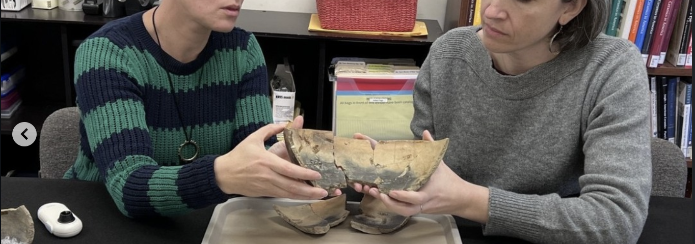 Elizabeth Bollwerk in striped sweater and Lindsay Bloch in gray sweater, sitting at a table and holding together a mending colonoware bowl over a tray, with other colonoware vessels in foreground.