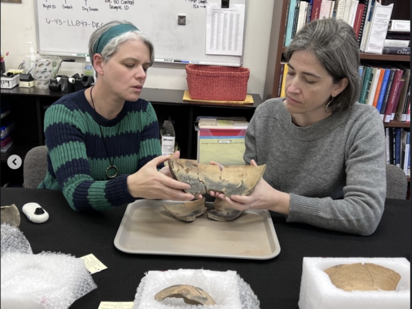 Elizabeth Bollwerk in striped sweater and Lindsay Bloch in gray sweater, sitting at a table and holding together a mending colonoware bowl over a tray, with other colonoware vessels in foreground.